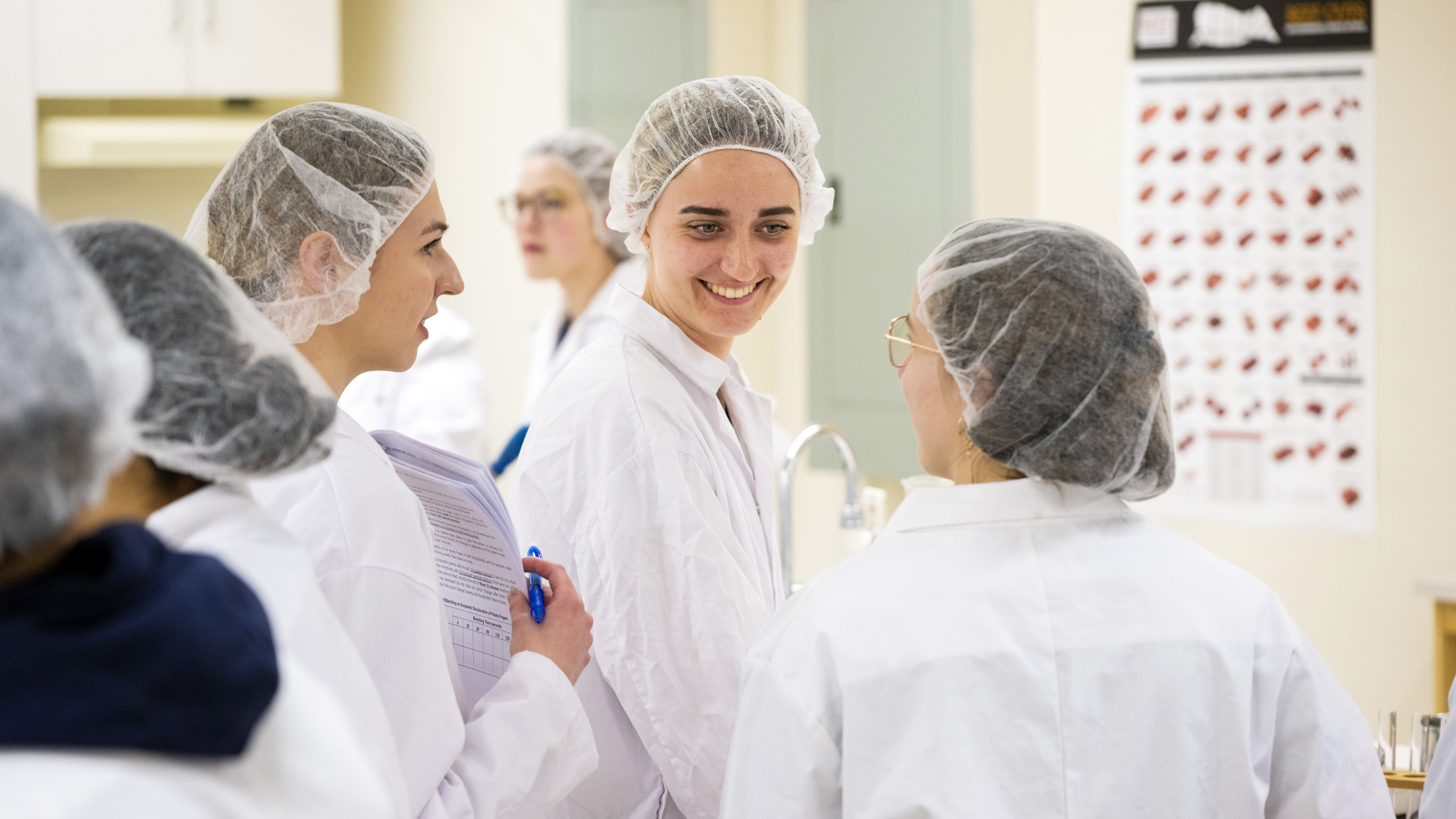 Students speak to one another in a lab setting wearing hair nets and lab coats
