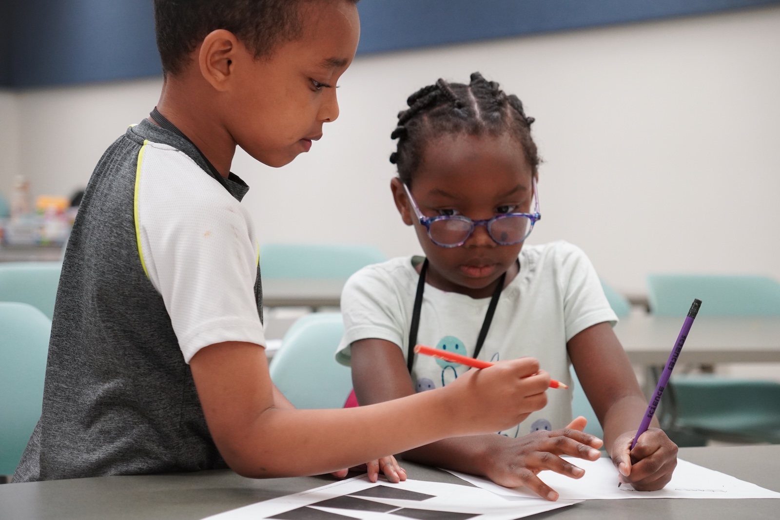 A group of six diverse children, wearing casual clothing and backpacks, pose in a classroom setting. Some wear name tags, and one child holds a small plastic bag while another gestures towards the camera. A hot plate sits on the table in front of them. The children appear to be engaged in a hands-on learning activity.